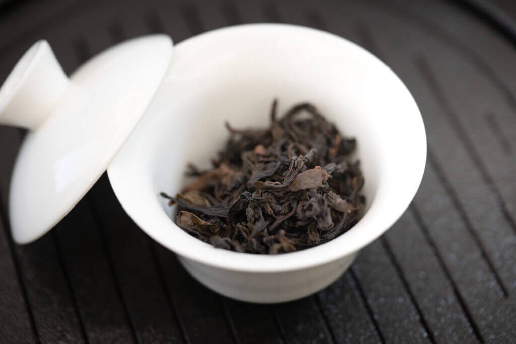Dried Tea Leaves in a White Porcelain Gaiwan with Lid Slightly Opened on a Dark Wooden Surface